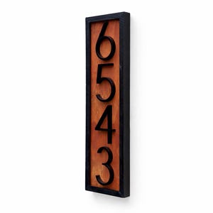 May include: Vertical wooden address sign with the numbers 6543 in black. The numbers are raised against a reddish-brown wooden background, framed by a black border. The sign is mounted on a white wall.