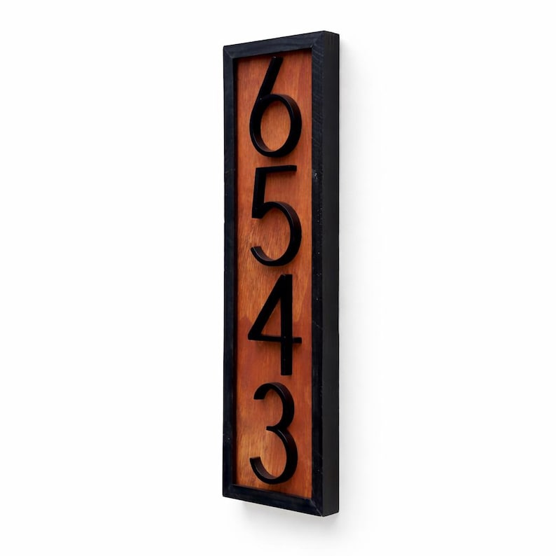 May include: Vertical wooden address sign with the numbers 6543 in black. The numbers are raised against a reddish-brown wooden background, framed by a black border. The sign is mounted on a white wall.