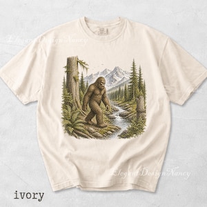 May include: Ivory-colored t-shirt featuring a detailed illustration of a Bigfoot walking near a stream in a forest, with mountains in the background. The text "ivory" is printed at the bottom of the shirt.