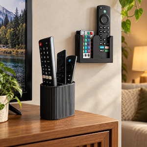 Op de afbeelding: Een zwarte, geribbelde, cilindervormige afstandsbedieningshouder staat op een houten oppervlak, terwijl een bijpassende zwarte, rechthoekige wandorganizer een meerkleurige led-afstandsbediening en een zwarte streaming-afstandsbediening bevat.