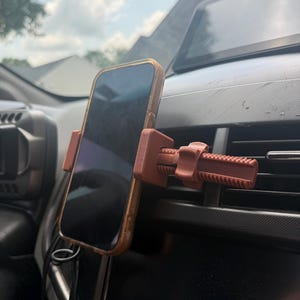 May include: A matte terracotta-colored plastic phone mount attached to a vehicle air vent. The mount features a horizontal adjustable screw mechanism with a threaded rod and a tightening nut to secure a smartphone in a vertical position.