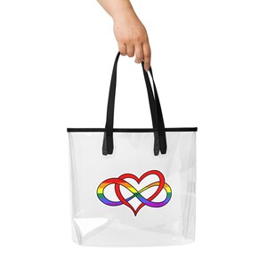 May include: Clear tote bag with black handles and trim. The bag features a rainbow-colored infinity heart design. The bag is held by a hand against a white background.