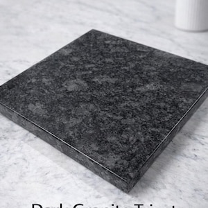 May include: A dark granite trivet made of solid stone. The square trivet has a speckled, dark gray appearance and is set against a light gray and white background. The text "Dark Granite Trivet" and "Solid Stone" are visible.