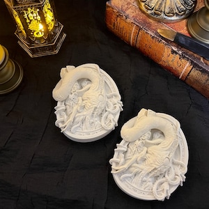 Puede incluir: Dos placas decorativas ovaladas de color blanco que presentan una escultura en relieve detallada de un dragón con un cuerpo largo y sinuoso y escamas texturizadas. El dragón se muestra en posición enroscada dentro del marco ovalado, con tallas intrincadas.