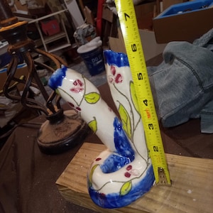 May include: A white ceramic vase with blue accents and floral designs, measuring approximately 7 inches tall. The vase features red flowers and green leaves, with a blue rim and base. It is placed on a wooden surface.