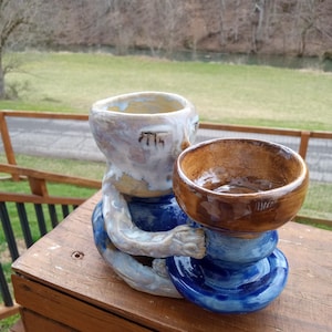 May include: A ceramic sculpture of a figure holding a bowl. The figure has a blue base and arms, with a white and brown head and upper body. The bowl it holds is brown. The sculpture is on a wooden surface.