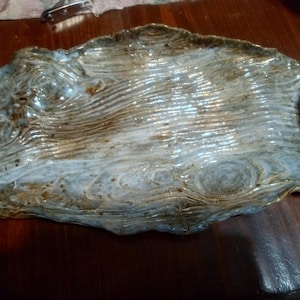 May include: A handcrafted ceramic serving tray with a unique, organic shape. The tray features a textured surface with a wood-grain pattern in shades of brown, white, and blue. The edges are uneven, adding to its rustic charm.