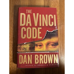 Pode incluir: Um livro de capa dura intitulado The Da Vinci Code de Dan Brown. A capa vermelha apresenta letras douradas e uma secção rasgada que revela uma parte da pintura da Mona Lisa com escrita à mão. O texto inclui A Novel e Author of Angels & Demons.