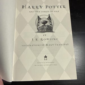 May include: The title page of Harry Potter and the Goblet of Fire by J.K. Rowling, illustrated by Mary GrandPré. The page features a light gray diamond pattern background, a central illustration of a flaming goblet, and the Arthur A. Levine Books logo.