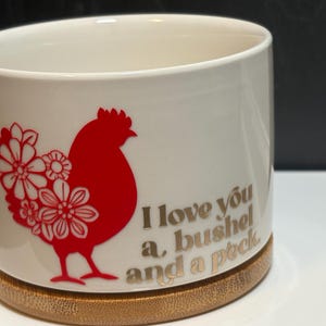 May include: A white ceramic cylindrical planter with a bamboo base. The front features a red silhouette of a chicken decorated with white floral patterns and the gold-colored text "I love you a bushel and a peck" printed to the right of the chicken.