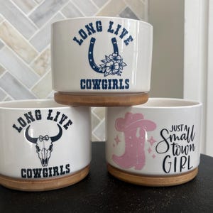 May include: Three white ceramic cylindrical planters with bamboo bases. The top planter features a blue horseshoe and flower graphic with the text LONG LIVE COWGIRLS. The bottom left shows a bull skull and LONG LIVE COWGIRLS. The bottom right shows a pink cowboy boot and the text JUST A SMALL TOWN GIRL.