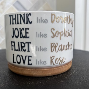 May include: A white ceramic cylindrical planter with a bamboo base. The text reads: THINK like Dorothy, JOKE like Sophia, FLIRT like Blanche, LOVE like Rose. The words THINK, JOKE, FLIRT, and LOVE are in black, while the names are in gold.