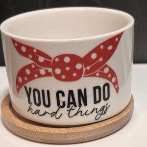 May include: White ceramic mug with a red polka-dot bow design and the text "YOU CAN DO hard things" in black. The mug sits on a round, light brown bamboo coaster. The mug is cylindrical and has a wide opening.