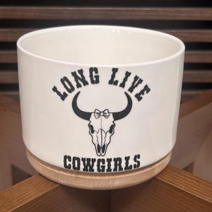 May include: A white ceramic planter with a cylindrical shape and a light wood base. The front features a black graphic of a bull skull with a small bow between the horns, surrounded by the black text "LONG LIVE COWGIRLS" in a western font.