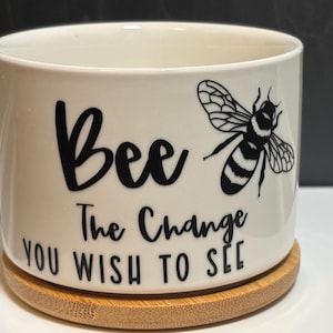 May include: A white ceramic planter with the words "Bee The Change You Wish To See" in black script. A black and white bee graphic is on the right side. The planter sits on a round, natural-colored bamboo coaster.
