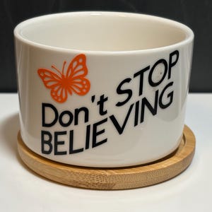 May include: A white ceramic planter with a cylindrical shape, featuring a bright orange butterfly graphic and the black text "Don't Stop BELIEVING" printed on the front. The planter rests on a circular, light-colored bamboo wood base.
