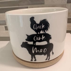 May include: A white cylindrical ceramic planter with a light wood base. The front features a black graphic of a rooster, a pig, and a cow stacked vertically. The text Cluck is on the rooster, Oink is on the pig, and Moo is on the cow.