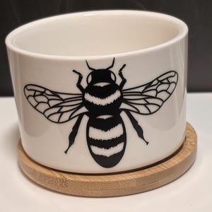 May include: White ceramic planter with a black and white bee illustration. The planter sits on a round, natural bamboo coaster. The planter has a slanted design, and the bee graphic is centered on the front.