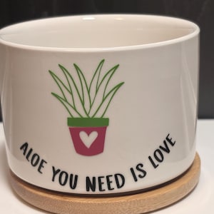 May include: White ceramic planter with a green aloe vera plant illustration in a pink pot with a white heart. The planter has the text "Aloe You Need Is Love" in black. It sits on a round, natural wood coaster.