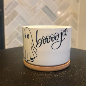May include: A white cylindrical ceramic planter featuring a black line-art illustration of a ghost with eyelashes and the black cursive text "booojee" printed on the side. The planter sits on a circular light brown wooden drip tray base.