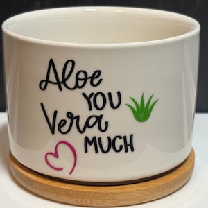 May include: A white cylindrical ceramic planter with a bamboo saucer base. The front features black text that reads Aloe YOU Vera MUCH, accompanied by a small green aloe plant graphic and a pink heart outline printed on the smooth surface.