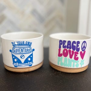 May include: Two white ceramic cylindrical planters with bamboo bases. The left planter features a blue graphic of a van with the text BE YOUR OWN ADVENTURE. The right planter displays the text PEACE LOVE PLANTS in purple, pink, and light green.