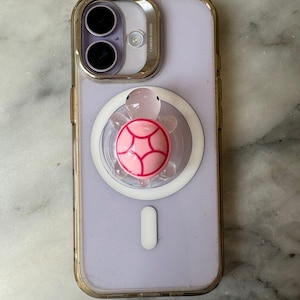 May include: A light purple iPhone with a clear case and a gold-colored trim. A pink turtle phone grip is attached to the back. The turtle grip has a pink shell with red accents and a clear head.