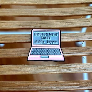 May include: A light pink enamel pin shaped like an open laptop computer. The screen displays the text 'DOCUMENT IT OR IT didn't happen' in black lettering against a light blue background. The keyboard area is dark gray with a light pink frame.