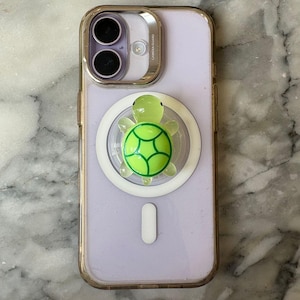 May include: A light purple smartphone with a clear case and a green turtle phone grip. The turtle grip has a green shell design and is attached to a white circular magnetic base.