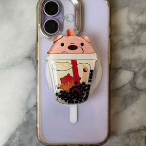 May include: A light purple smartphone with a gold-colored case and a cartoon-style pop socket. The pop socket features a bear character in a boba tea cup with black tapioca pearls and a red straw.