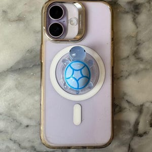 May include: A light purple iPhone with a clear case and a turtle-shaped phone grip. The grip is blue and white, and the phone has a gold-colored camera guard.