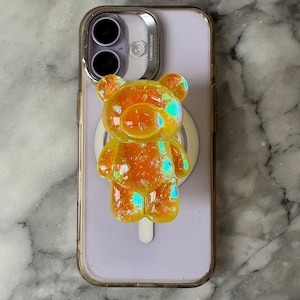 May include: A clear phone case with a light purple phone inside. An orange, sparkly bear-shaped phone grip is attached. The bear grip has iridescent glitter and a white circular grip.