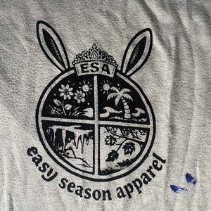 May include: A light gray t-shirt featuring a black graphic. The graphic includes a circular design divided into four sections, each with a different illustration: flowers, palm trees, a waterfall, and leaves. The text "ESA" is at the top, and "easy season apparel" is around the circle.
