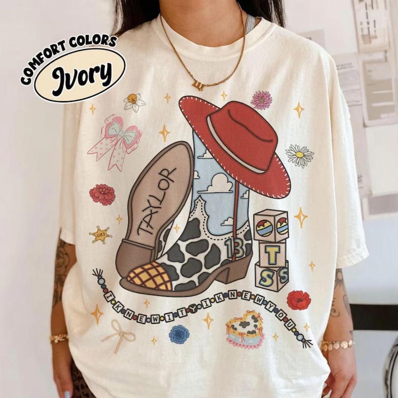 May include: An ivory t-shirt featuring a graphic print of a cowboy boot with a cow print pattern, a red cowboy hat, and the name Taylor. The design includes blocks with the letter T, a beaded string, flowers, and stars. Text reads COMFORT COLORS IVORY.