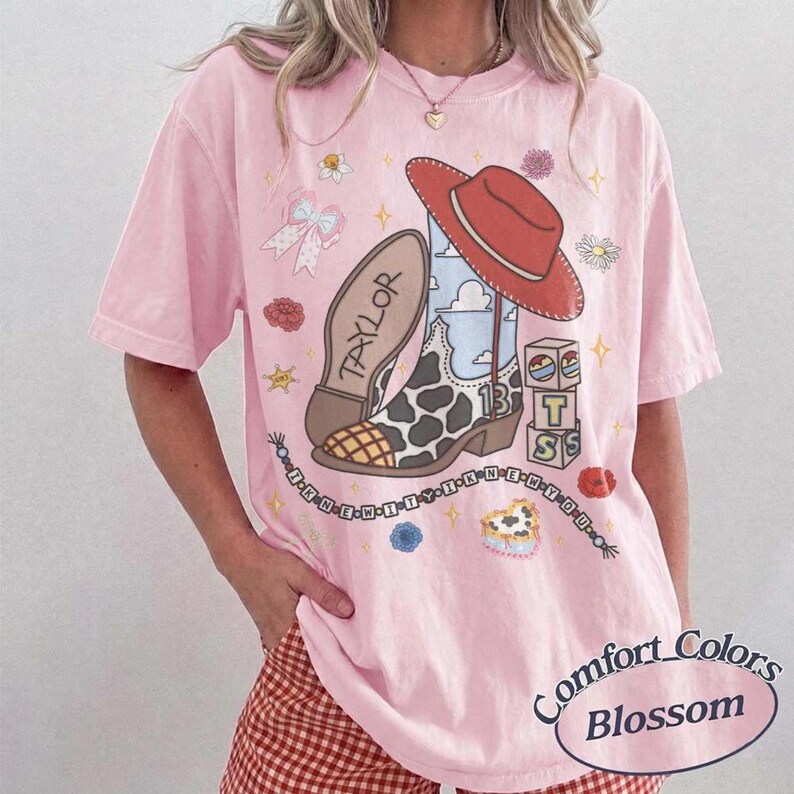 May include: A light pink short-sleeve t-shirt featuring a graphic print of a cowboy boot with a cow print pattern, a red cowboy hat, a bow, flowers, and blocks with the text "TAYLOR" and "13". A string of beads spells out "I KNEW YOU WERE TROUBLE".