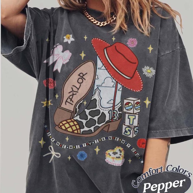May include: A charcoal gray t-shirt featuring a graphic print of a cow-print cowboy boot, a red cowboy hat, and the name Taylor. The design includes blocks with the letters BTS, a heart-shaped cake, bows, flowers, and the text I KNEW IT Y I KNEW YOU.