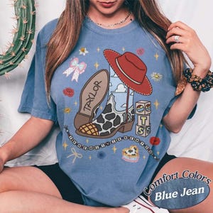 May include: A blue short-sleeve t-shirt featuring a graphic print of a cow-print cowboy boot, a red cowboy hat, and blocks spelling T-S. The boot is labeled with the name Taylor and the number 13, surrounded by stars, flowers, and a beaded string.