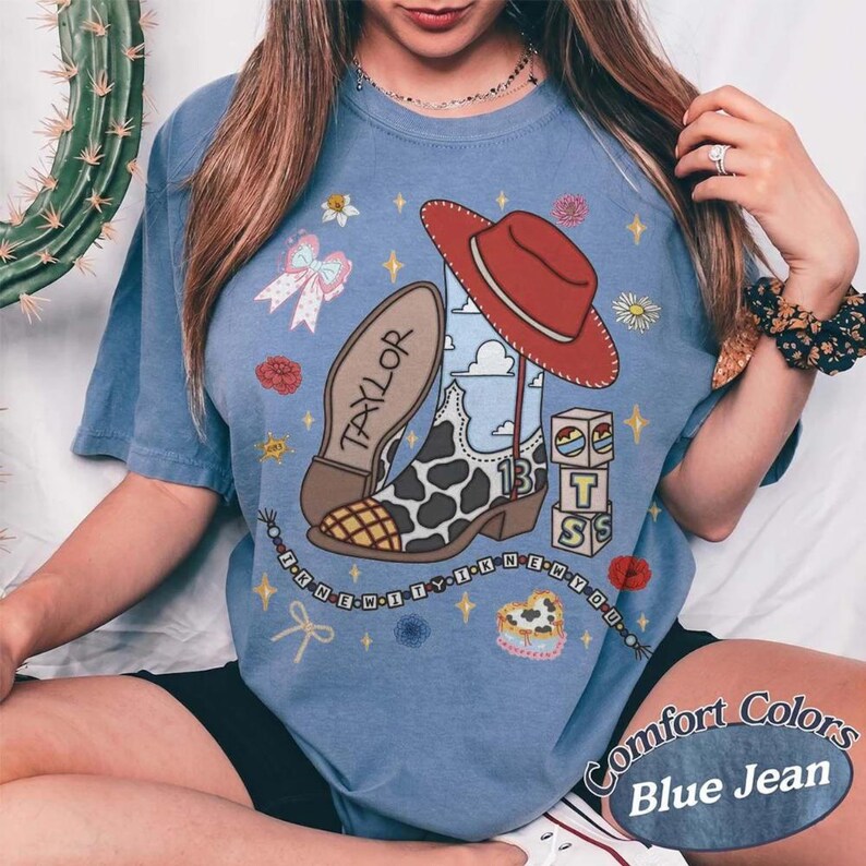 May include: A blue short-sleeve t-shirt featuring a graphic print of a cow-print cowboy boot, a red cowboy hat, and blocks spelling T-S. The boot is labeled with the name Taylor and the number 13, surrounded by stars, flowers, and a beaded string.