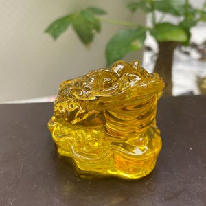 May include: A translucent amber-colored glass figurine of a three-legged toad, known as a Jin Chan, depicted with a textured, bumpy back and sitting atop a base of stacked coins. The sculpture features a glossy finish and reflective surfaces.