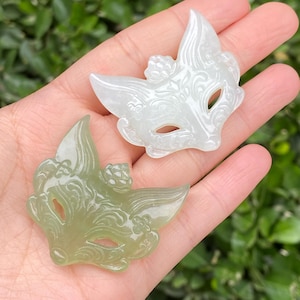 May include: Two decorative fox masks. One is a pale green color, and the other is white. Both masks have eye holes and are carved with floral and swirl designs. The masks are made of a translucent material.