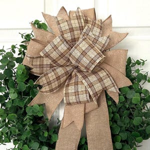 May include: A circular wreath composed of dense green eucalyptus leaves, featuring a large decorative bow at the top. The bow is crafted from a combination of solid tan burlap ribbon and a tan, brown, and white plaid patterned ribbon with long tails.