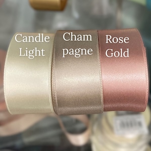 May include: Three rolls of smooth satin ribbon arranged side by side. The left roll is labeled Candle Light, the center roll is labeled Champagne, and the right roll is labeled Rose Gold. Each ribbon features a solid color and a subtle sheen.