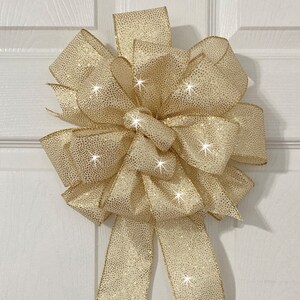 May include: A large gold glitter bow with a long tail. The bow is tied with a double knot and has a shimmery finish.