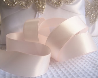 May include: A close-up of a soft, ivory satin ribbon. The ribbon is folded and arranged on a white textured surface.