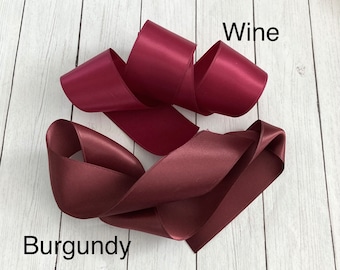 May include: Two rolls of satin ribbon, one in burgundy and one in wine. The ribbon is folded and arranged on a white wooden surface.