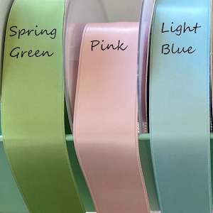 May include: Spools of satin ribbon in spring green, pink, and light blue. The ribbon is displayed with the color names written in black script. The ribbon is likely used for crafts, gift wrapping, or decorative purposes.