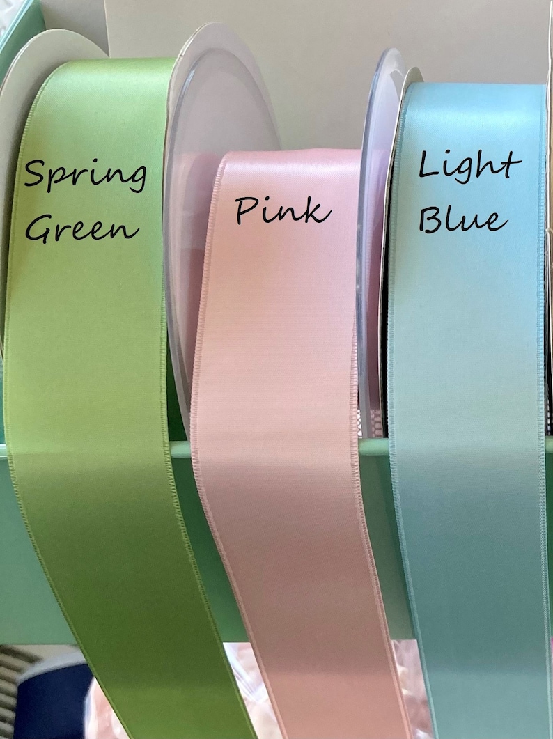 May include: Spools of satin ribbon in spring green, pink, and light blue. The ribbon is displayed with the color names written in black script. The ribbon is likely used for crafts, gift wrapping, or decorative purposes.