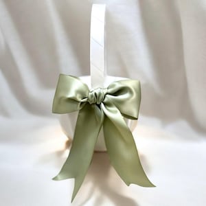 May include: A white basket with a handle, adorned with a large, sage green satin bow. The bow is tied in the center and has long, flowing tails. The basket is likely for decorative purposes.