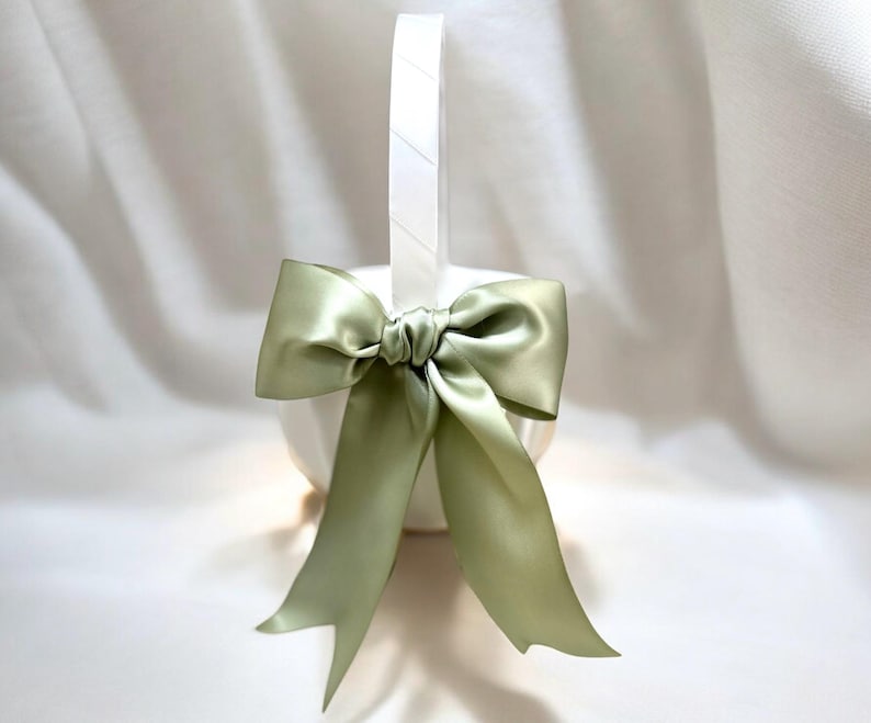 May include: A white basket with a handle, adorned with a large, sage green satin bow. The bow is tied in the center and has long, flowing tails. The basket is likely for decorative purposes.