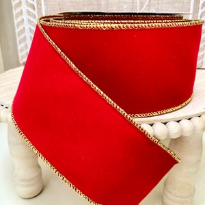 May include: Red velvet ribbon with a gold trim. The ribbon is rolled up and is sitting on a white pedestal.